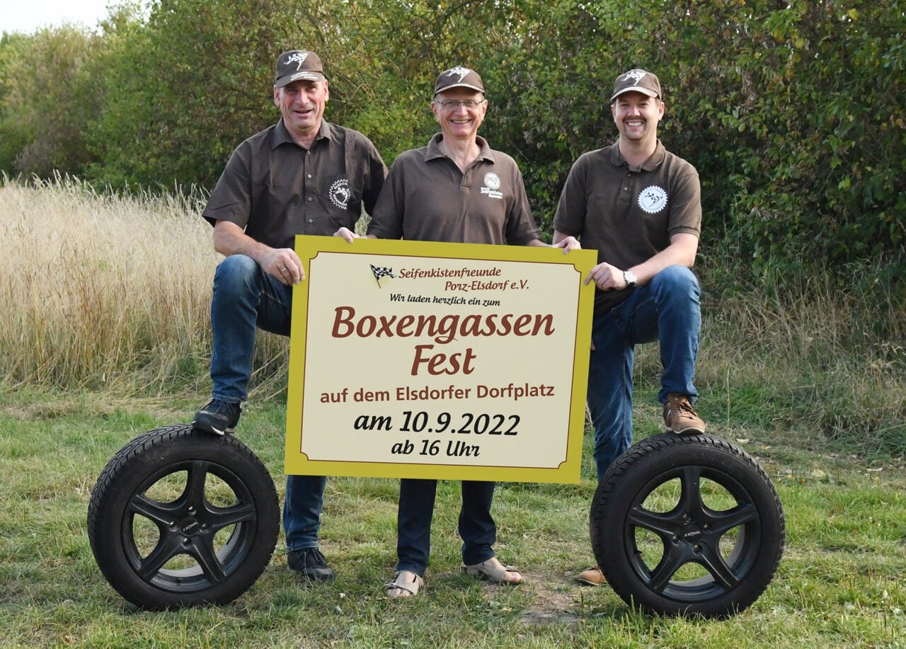 Drei Männer stehen neben einem Schild mit der Aufschrift Boxengassenfest.